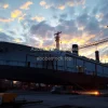 Shipbuilding yard with massive hull under construction cranes and welders working sunset lighting realistic